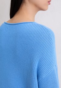 Sweatshirt en tricot bleu avec une texture côtelée, présentant un large décolleté et une coupe ample, mettant en avant un tissu doux et lisse couvrant l'épaule.