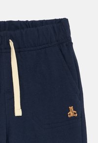 Pantaloni felpati blu navy con cintura elastica, cordoncino crema e un piccolo orsetto ricamato sulla tasca anteriore. Tessuto morbido.