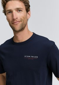 Tamsiai mėlynas medvilninis marškinėlis su apvalia iškirpte, priekyje esantis baltas ir raudonas "TOM TAILOR" logotipas, lygus paviršius, trumpomis rankovėmis.
