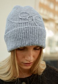 Grijze gebreide muts met een geribbelde structuur en geborduurde strikdetail aan de voorkant, nauwsluitend ontwerp, zacht materiaal en opgerolde rand.