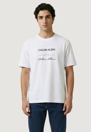 Hombre de cabello oscuro que lleva una camiseta blanca de Calvin Klein y vaqueros azules, de pie frente a un fondo claro y liso.