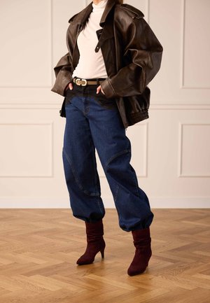 Femme portant une veste en cuir marron surdimensionnée, un col roulé blanc, un jean bleu foncé ample, une ceinture noire et des bottes à talons rouge foncé, debout sur un sol en bois.