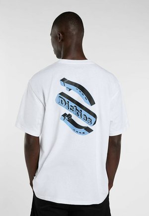 Uomo che indossa una maglietta bianca con logo 3D "Dickies" blu e nero sulla schiena, in piedi davanti a uno sfondo chiaro e uniforme.