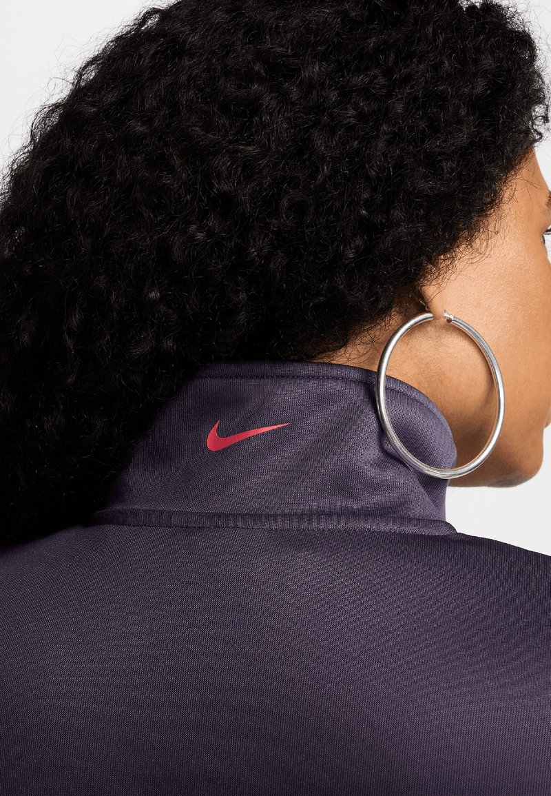 Chaqueta de Nike en tela de color púrpura oscuro con un cuello alto. Presenta un logo de swoosh rojo y una persona que lleva un gran pendiente de aro plateado.
