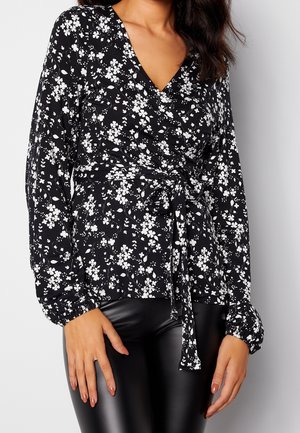 Schwarze Wickelbluse mit langen Ärmeln und weißem Blumenmuster sowie gebundenem Taillenband, kombiniert mit schwarzen Lederhosen an einer Frau.