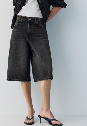 Oseba, oblečena v črne široke kavbojke z visokim pasom, belo majico, črno jakno in črne sandale s peto, stoji ob enobarvni steni.