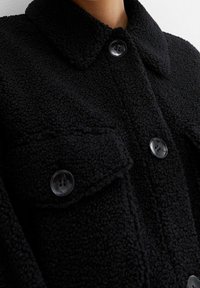 Chaqueta negra de felpa con cuello tipo solapa, dos bolsillos en el pecho y botones redondos negros, hecha de una suave tela texturizada.