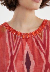 Haut à motifs rouges avec un large décolleté orné d'une bordure en perles rouges vives, complété par un tissu doux et texturé.