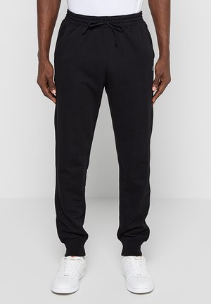Uomo che indossa pantaloni jogger neri con vita elastica e coulisse, abbinati a sneaker bianche e camicia bianca.