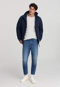 Giacca imbottita blu navy con cappuccio, maglietta grigia a girocollo, jeans azzurri a gamba stretta, sneakers bianche. Design semplice, texture liscia.