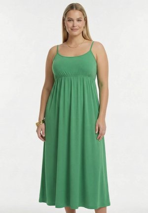 Kvinde med langt blondt hår iført en ærmeløs, grøn kjole, der går til anklerne, med tynde stropper og gyldne armbånd på venstre håndled.