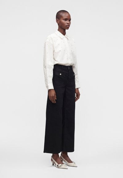 Femme portant une chemise blanche à manches longues avec de petits motifs, un pantalon noir à jambes larges, et des chaussures blanches à petits talons avec des boucles.