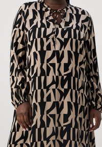 Personne portant une robe beige avec d’audacieux motifs géométriques abstraits noirs, manches longues et nœud à l’encolure. Mains détendues le long du corps.