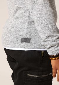 Grijze, gestructureerde trui met een label aan de zijkant, gecombineerd met zwarte broek met ritszakken en een contrasterende witte zoom.