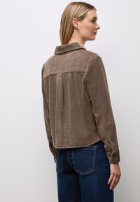 Camisa de pana marrón con cuello, espalda con pliegues y puños abotonados, combinada con jeans azules. La tela texturizada realza el interés visual.