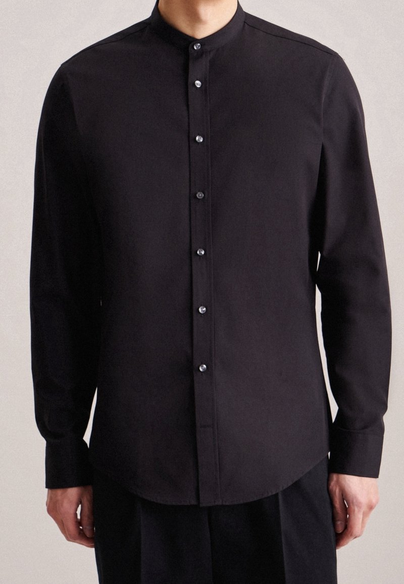 Homme portant une chemise noire à manches longues avec un col mandarin et des boutons sur le devant, debout les bras détendus le long du corps.