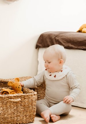 Kind met kort licht haar, gekleed in een beige outfit met lange mouwen en een witte slab met rode accenten, zittend naast gevlochten manden en een teddybeer vasthoudend.
