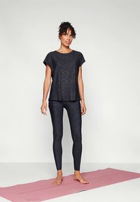 Fekete atlétikai póló rövid ujjakkal és finom mintázattal, fekete leggingsszel, melynek mintázata texturált, egy rózsaszín jógaszőnyegen.