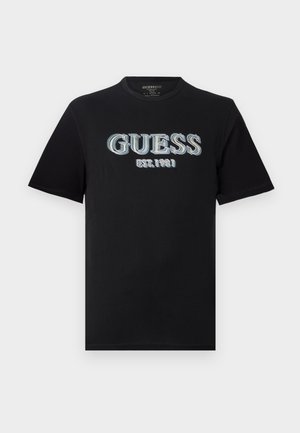Black cotton T-shirt featuring a prominent blue and white embroidered "GUESS" logo with "EST. 1981" underneath. Short sleeves and round neck.
