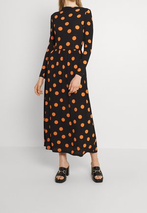 Femme portant une robe midi noire à manches longues avec des pois orange vif et des sandales à talons carrés ouvertes noires, debout sur un sol gris.