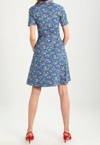 Kortärmad klänning i blått med blommönster, med en figursydd överdel och utsvängd kjol. Buren med röda klackar.