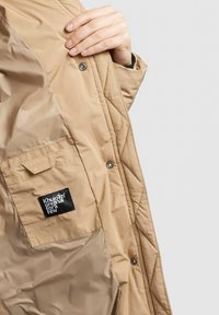 Veste beige matelassée, avec fermeture à pression, présentée avec une poche plaquée et un label noir. Texture lisse, design minimaliste.
