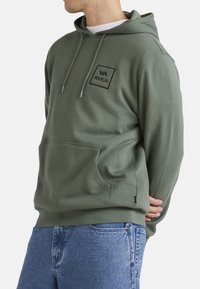 Olive green hoodie with a drawstring hood, front pocket, and a square logo patch on the chest. Made from soft, textured fabric.