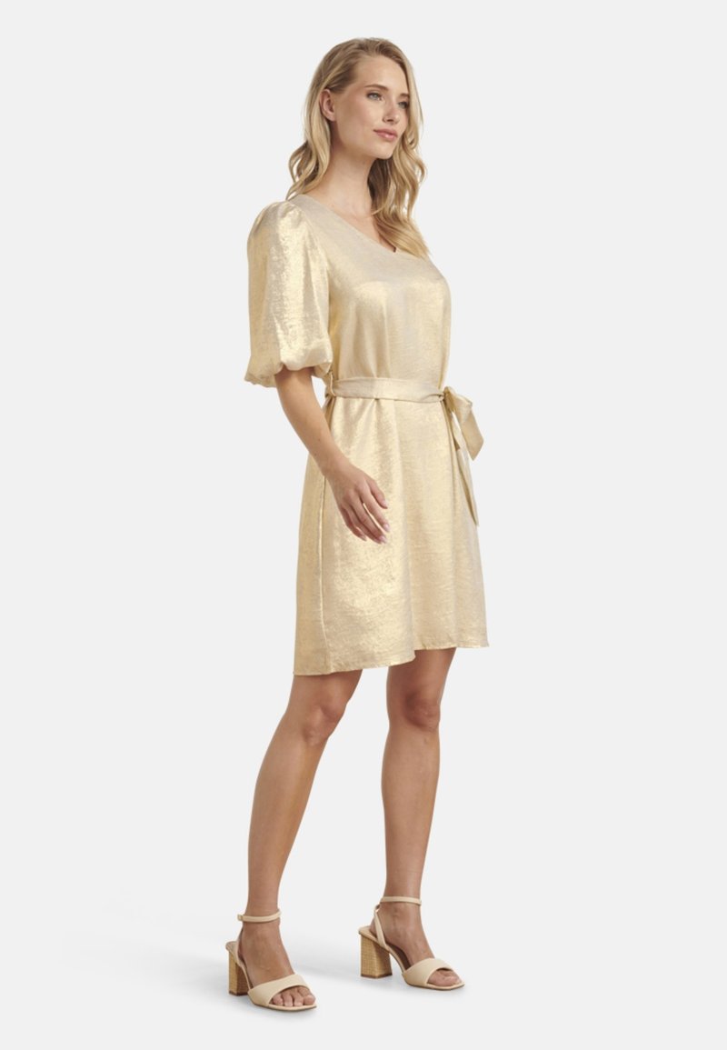 Goldfarbiges, kurzärmeliges, mit Gürtel versehenes Kleid aus glänzendem Stoff mit lockerer Passform und knielanger Länge. Begleitet von hellen High Heels.