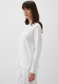 Maglietta bianca a maniche lunghe con vestibilità rilassata, fessure laterali e scollatura tonda, realizzata in tessuto liscio con un leggero lucido.