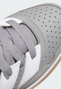 Šedá športová obuv s perforovaným syntetickým zvrškom, bielymi akcentmi, plochými šedými šnúrkami a textúrovanou gumenou podrážkou. Viditeľné detailné švy.