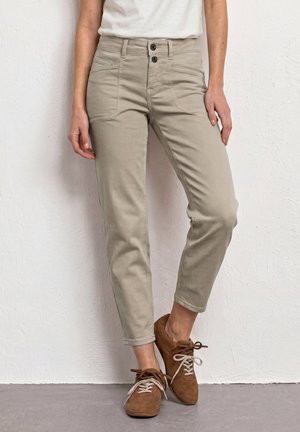 Vrouw draagt beige slim-fit broek met twee knopen vooraan en bruine veterschoenen, staand tegen een gestructureerde witte muur.