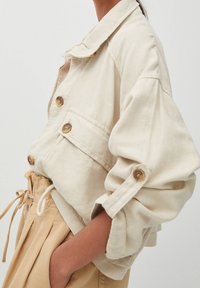Veste courte beige en tissu avec de grands boutons, col replié et détail de poche latérale. Caractéristiques de manches retroussées et coupe décontractée.