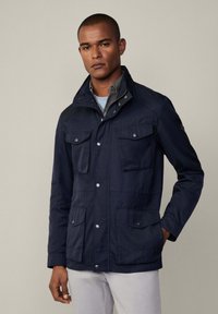 Chaqueta de color azul marino, con alto cuello, cuatro bolsillos exteriores, botones a presión y tejido texturizado; presenta un diseño en capas y una silueta ajustada.