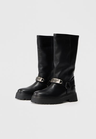 Botas de cuero negro hasta la rodilla con suela gruesa, hebilla metálica decorativa con diseño de ala y textura suave.