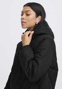Doudoune noire avec capuche, dotée d'une texture matelassée, de poignets élastiques et d'une poche latérale zippée. Fini lisse, design léger.