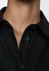 Camicia nera con bottoni, caratterizzata da un colletto aderente, bottoni visibili e un tessuto testurizzato. Focus sull'area del collo e sui dettagli della camicia.