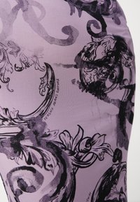 Fioletowa tkanina z czarnymi kwiatowymi i ozdobnymi wzorami, z napisem "Versace Jeans Couture" w dekoracyjnym stylu.