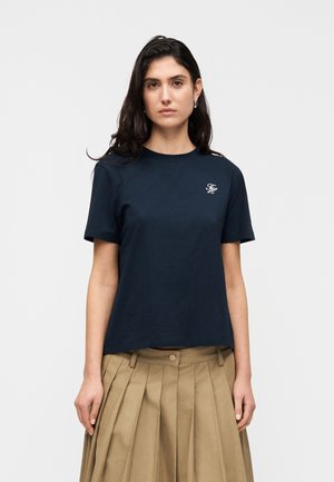 Žena tamne kose odjevena u tamnoplavu majicu s bijelim logom na prsima i bež plisiranu suknju, stoji ispred jednobojne pozadine.