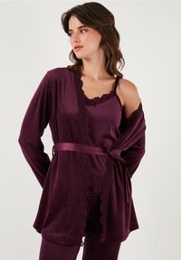 Young woman wearing dark purple velvet pajama set with lace trim, including a belted robe and camisole, looking to her left.