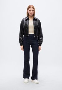 Μαύρο κοντό δερμάτινο μπουφάν με φερμουάρ μπροστά, φορεμένο πάνω από ένα μπεζ τοπ και σκουρόχρωμα παντελόνια με καμπάνα, συνδυασμένο με λευκά αθλητικά παπούτσια.