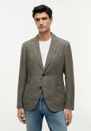 Mand iført en grå blazer over en hvid skjorte og blå jeans, stående mod en ensfarvet hvid baggrund, vendt fremad.