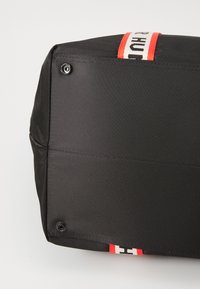 Bolsa de tela negra con superficie texturizada, que presenta acentos de hardware a rayas rojas y negras y pies de metal visibles en la parte inferior.