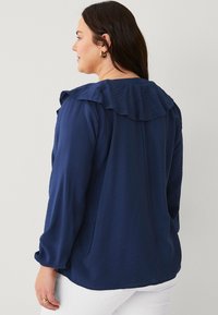 Blusa blu navy realizzata in tessuto testurizzato, con colletto arricciato e maniche lunghe. La vista posteriore mostra un orlo dritto e dettagli di cucitura sottili.