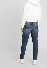 Jean en denim bleu foncé avec une jambe droite, présentant des détails effilochés et un ourlet effiloché. Associé à un hoodie en coton blanc.