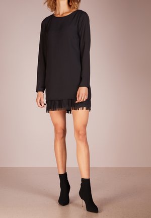 Femme portant une mini robe noire à manches longues avec un ourlet à franges, associée à des bottines à talons noirs, debout devant un fond uni.