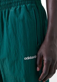 Calções leves verdes com cintura elástica, bolso lateral com fecho e logo da Adidas bordado na parte frontal esquerda. Tecido texturizado.