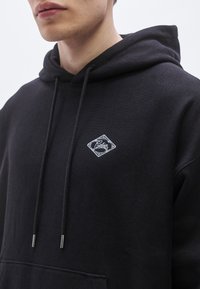 Čierna mikina s kapucňou vyrobená z mäkkej látky s bielym vyšívaným logom v tvare diamantového tvaru. Má šnúrky s kovovými koncovkami a predné vrecko.