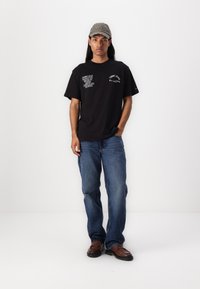 Sort bomulds t-shirt med hvid tekstgrafik. Personen har blå denimjeans og brune slip-on sko på, suppleret med en grå kasket.