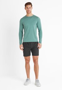 Man standing wearing a teal long-sleeve shirt, black shorts, and light gray athletic shoes against a white background.