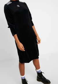 Svart sammet klänning med långa ärmar och hög halsringning, med tre vita ränder på axlarna och en Adidas-logotyp på bröstet.